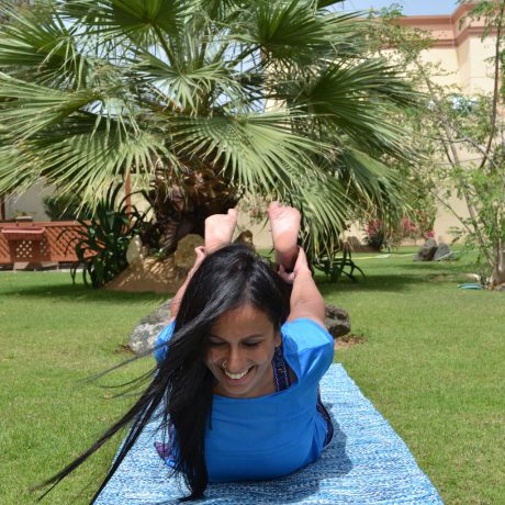 Hema Laxman, the owner of Flamingo Yoga & Wellness, practicing Dhanurasana (Bow Pose) on a natural cotton yoga mat in a lush green environment with palm trees in the background.